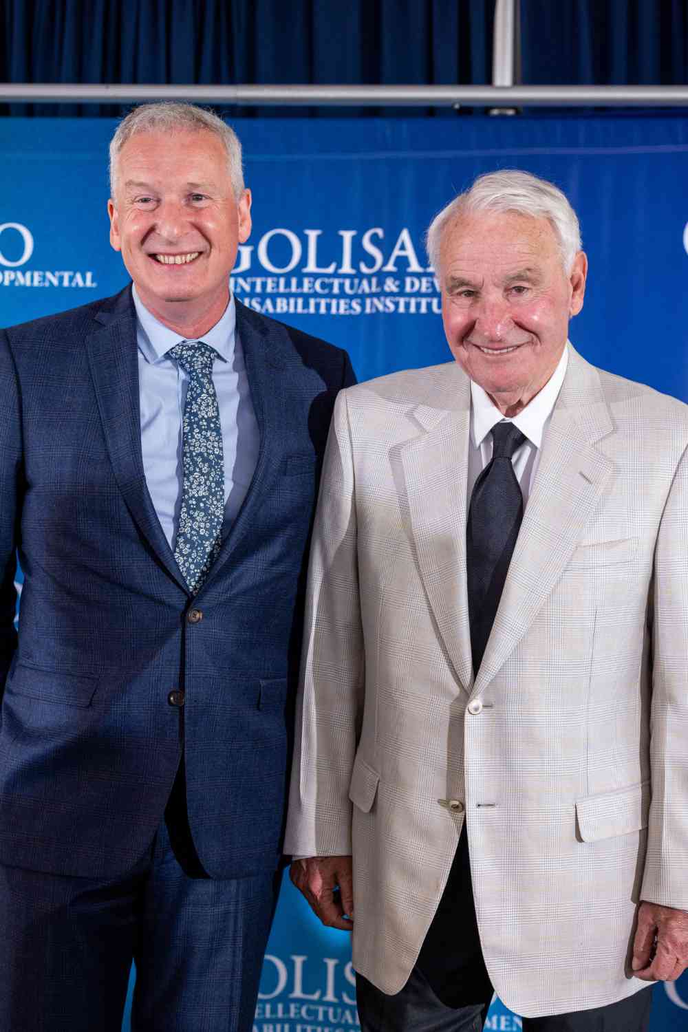 John Foxe and Tom Golisano in front of a blue backdrop featuring the University of Rochester logo for the Golisano Intellectual and Developmental Disabilities Institute. 