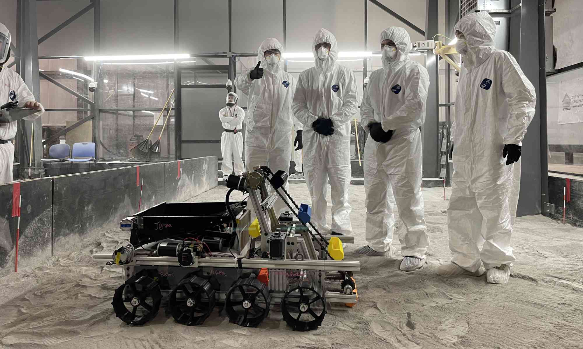 Four students in protective white gear pose with their lunar rover robot in a simulation area resembling the moon's terrain.