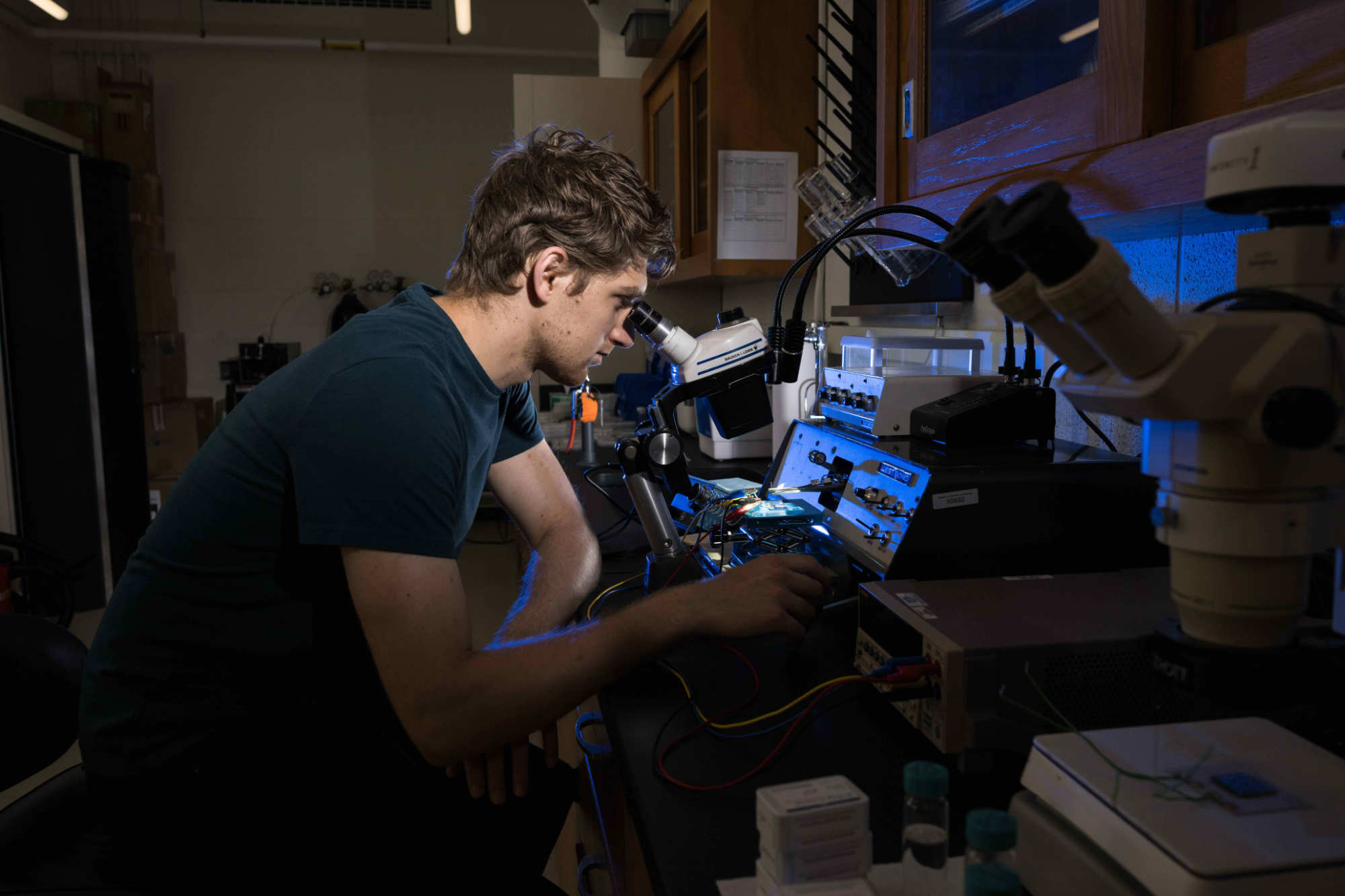 A graduate student doing nanoscale heat transfer mapping research peers into a microscope-like device while the screens and array in front of him are lit up with blue light.