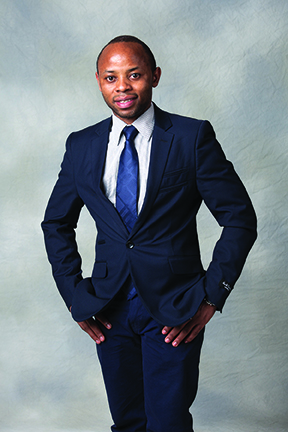 Bienfait Mugenza in blue suit and tie standing against gray background.