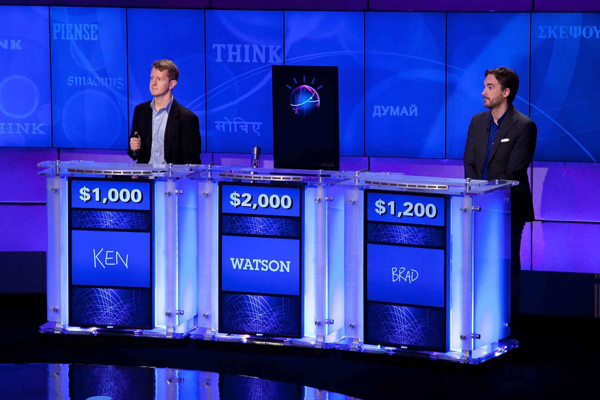 Jeopardy! gameshow contestants Ken Jennings, IBM's Watson computer, and Brad Rutter stand at a replica of the show's stage during a press conference.