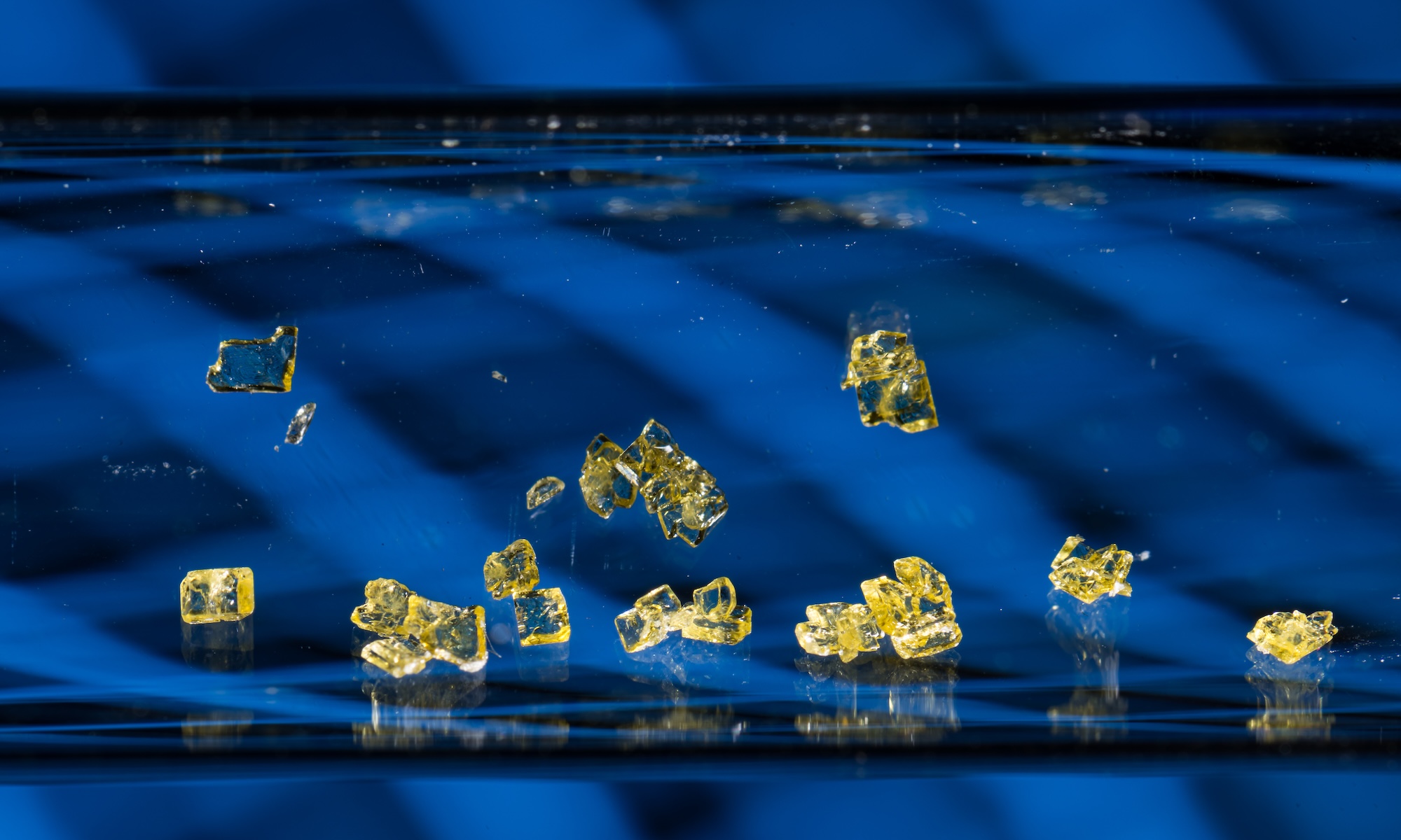 Closeup of yellow indoline crystals suspended in test tube against blue background.