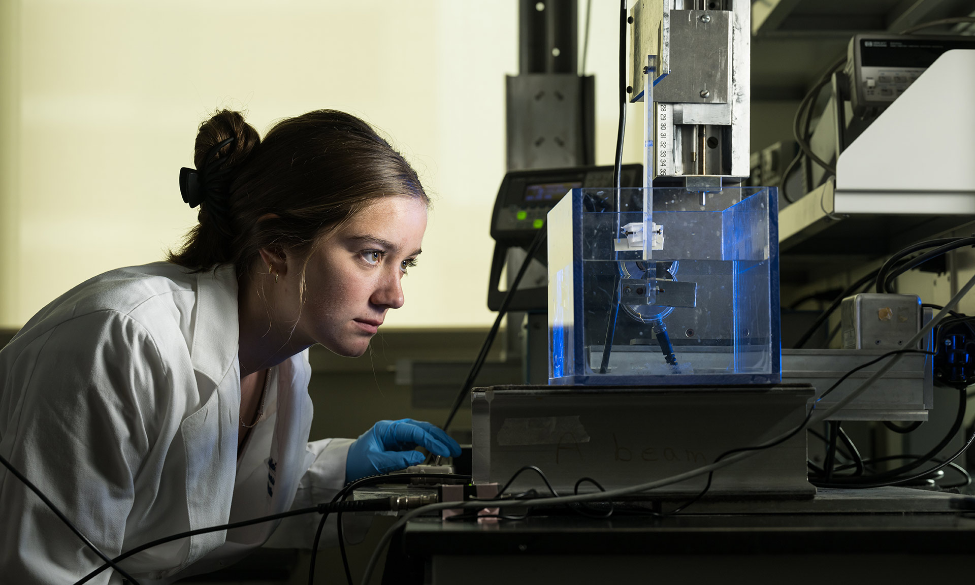 researchers in a white lab coat looks into brightly lit clear box reflected with blue light. 