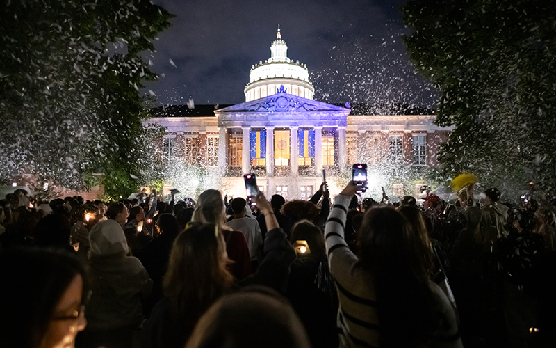 Students cheer and hold up phones to record in front of a lit-up Rush Rhees Library during Candlelight Ceremony
