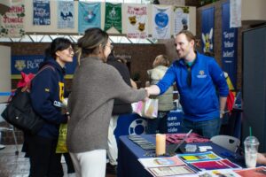 Individual wearing a blue U of R quarter zip hoodie tabling at a Wellness Fair smiling and greeting two students and shaking one of their hands. (Flourish Festival 2024)