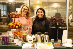 Two individuals smiling and standing behind a table while tabling about mindful eating, with coffee, tea, clementine oranges, and chocolate on the table in front of them. (Flourish Fest 2024)