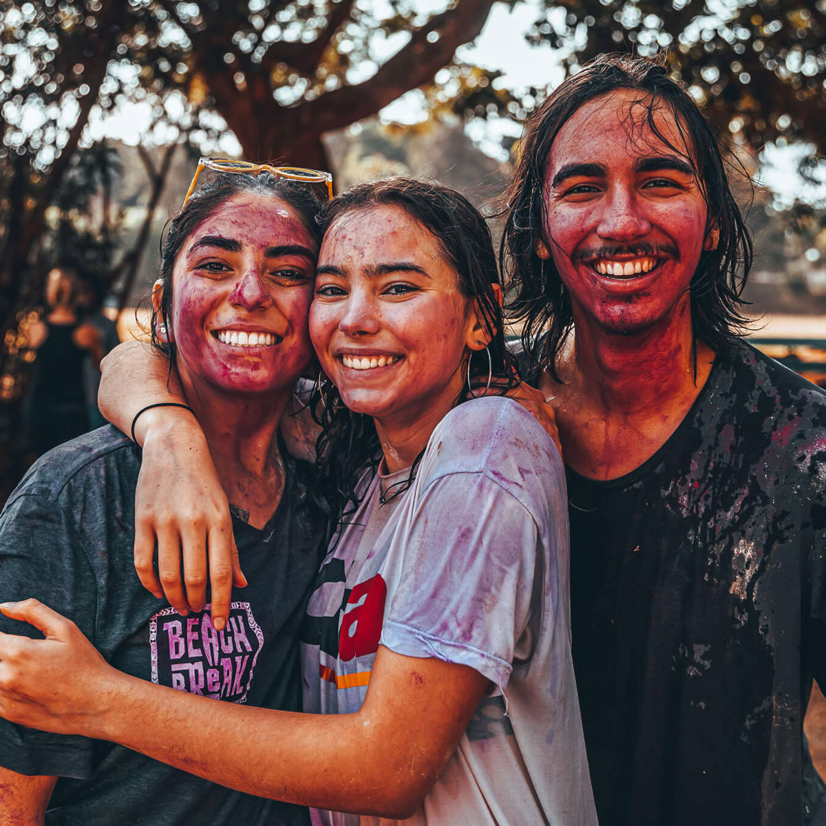 Three young people covered in colorful powder stand outdoors, smiling and hugging, with trees in the background.
