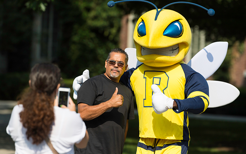 Photo of a woman taking a photo of a man posing with University of Rochester mascot, Rocky