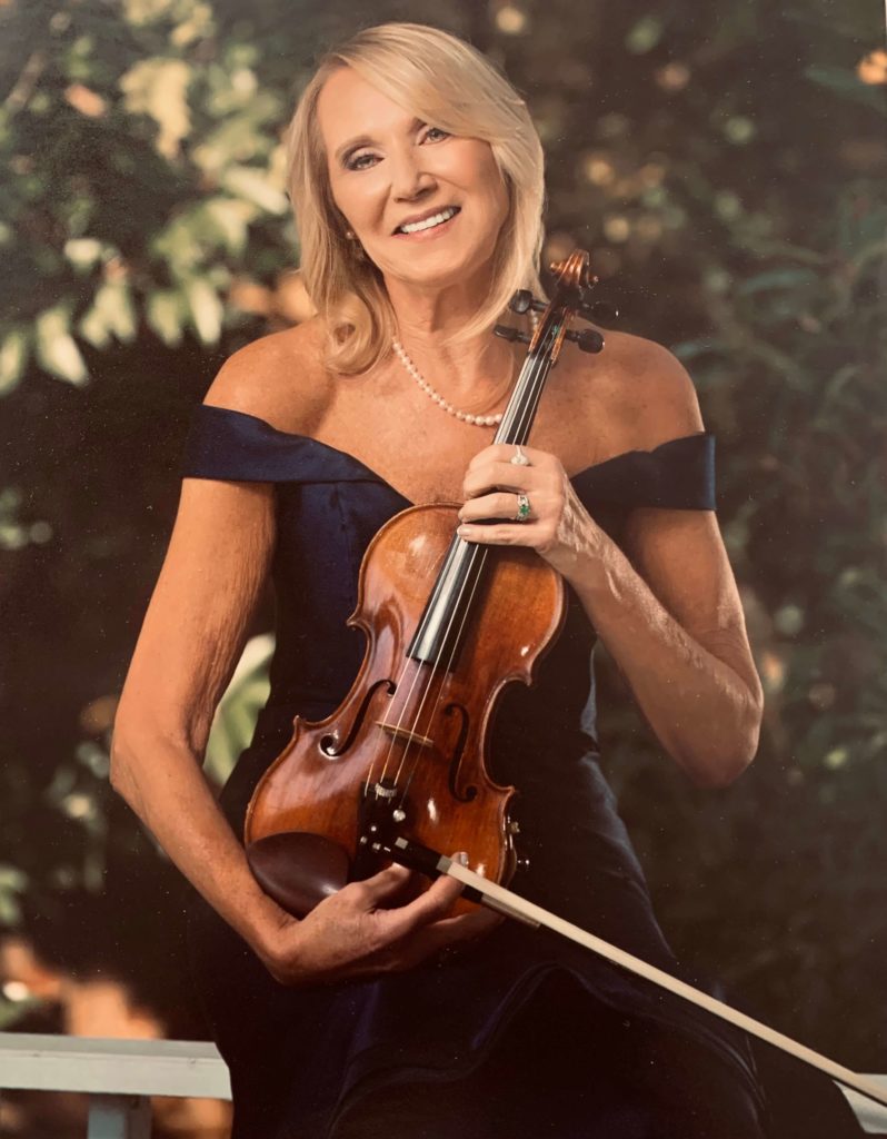 Photo of Eastman School of Music alumna Andrea Tolmich Shalhoub as she holds a violin while wearing a dark blue dress in a outdoor setting with trees in the background