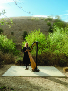Marry Lattimore posing with her harp in front of hill and bushes
