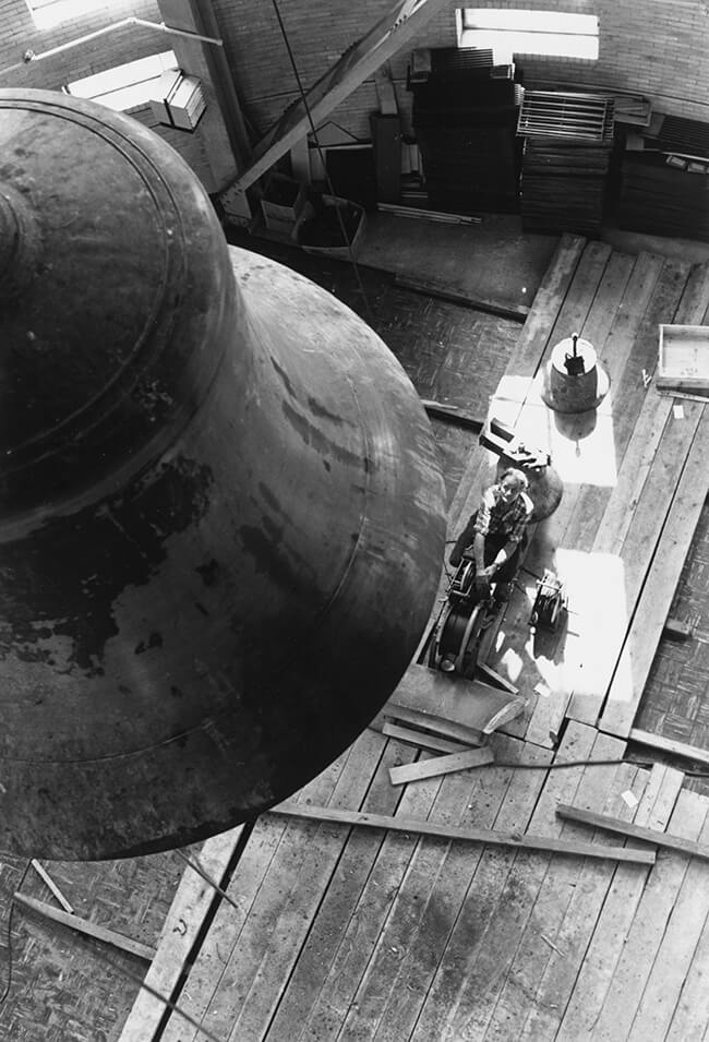 An Ariel black and white image of the Hopeman Memorial Carillon within the tower of Rush Rhees Library. A man is below it and looking upward