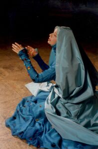 woman with long-sleeved blue dress and blue headdress sitting on stage with outstretched arms