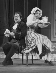 black and white photo of a man wearing a wig and coattails woman in a bonnet and long skirt fumbling a tray with a teapot