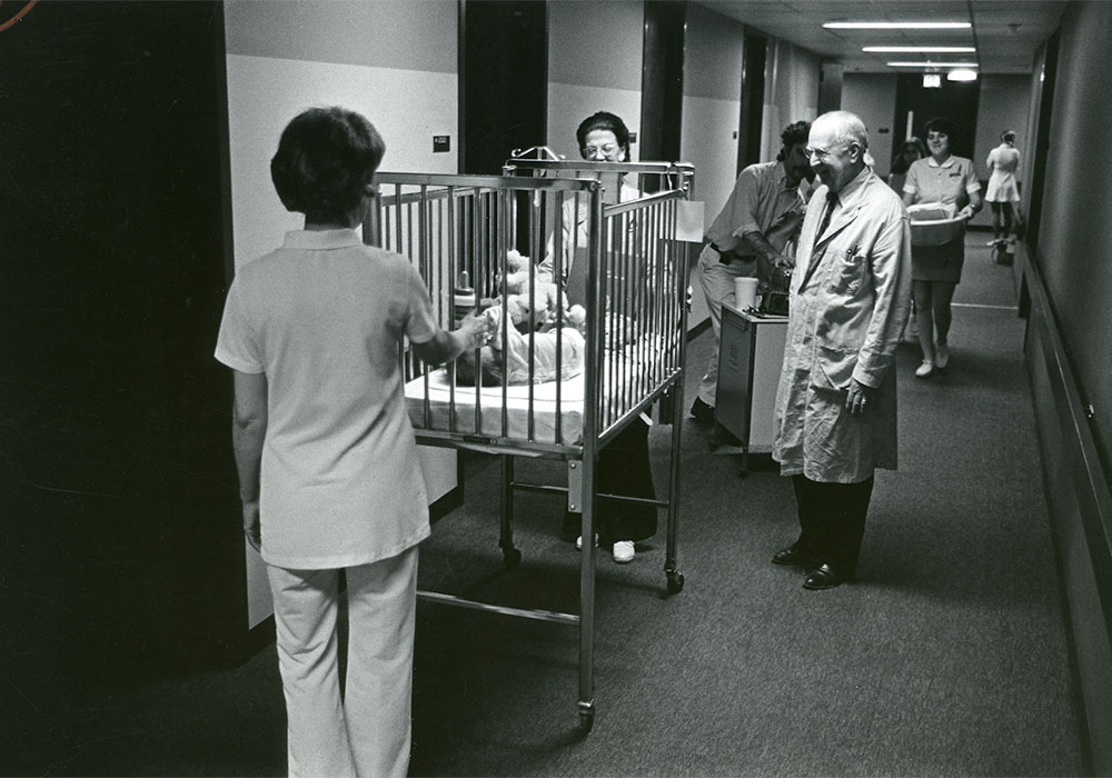 Doctor watching nurses move a baby in crib down a hall in Strong Memorial Hospital in 1975