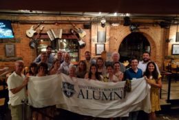 large group holding a rochester alumni banner at an event with brick wall behind them