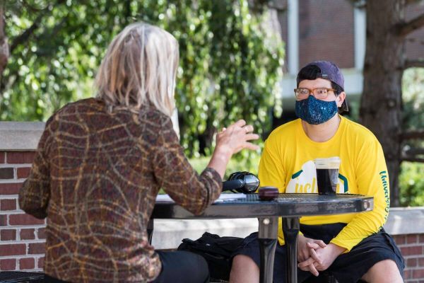 A student sitting at a bench outdoors talking with a teacher while wearing a face mask.
