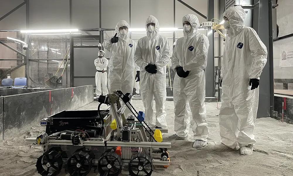 Four students in lab clean suits gather around a multi-wheeled robot on the ground.'