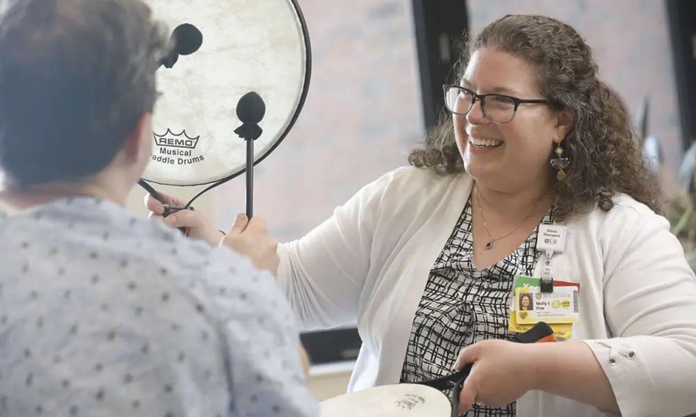 A music therapist smiles and holds paddle drums while a patient in a hospital gown seen from behind uses paddle sticks to beat the drums.