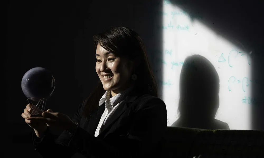 Niki Makajima smiles and looks at a replica moon sculpture while her shadow falls on a whiteboard with equations on it behind her.