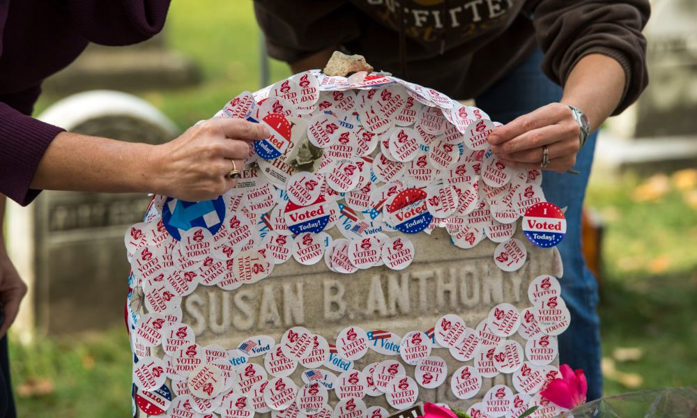 Gravestone of Susan B. Anthony covered with 'I Voted' stickers, with two hands adding more.