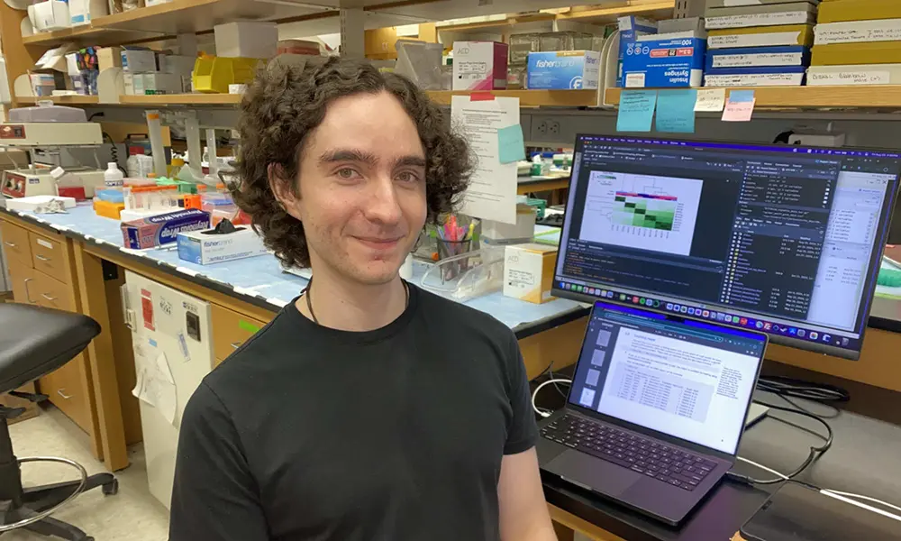 Alejandro Cruz Setzekorn smiles at the camera with a laptop and monitor in the background showing data science genomics research and figures.