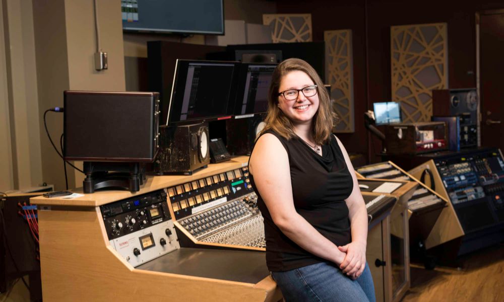 Woman standing in a recording studio with mixing console and audio equipment.'