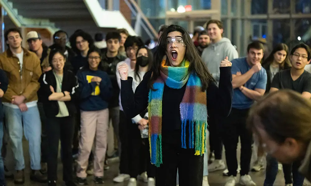 Student wearing protective goggles raises her fists in celebration and gives an expression of surprise as she stands in the Goergen Hall atrium surrounded by a crowd of students.'