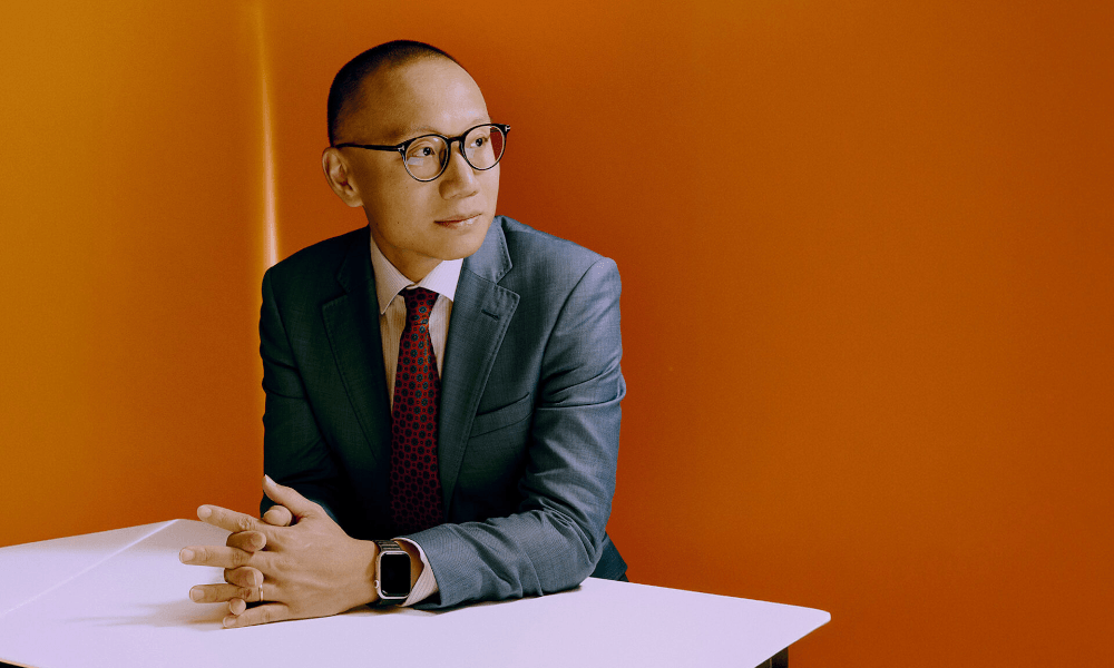 Dan Wang sits at a desk in front of an orange background with his fingers interlaced, looking away from the camera.