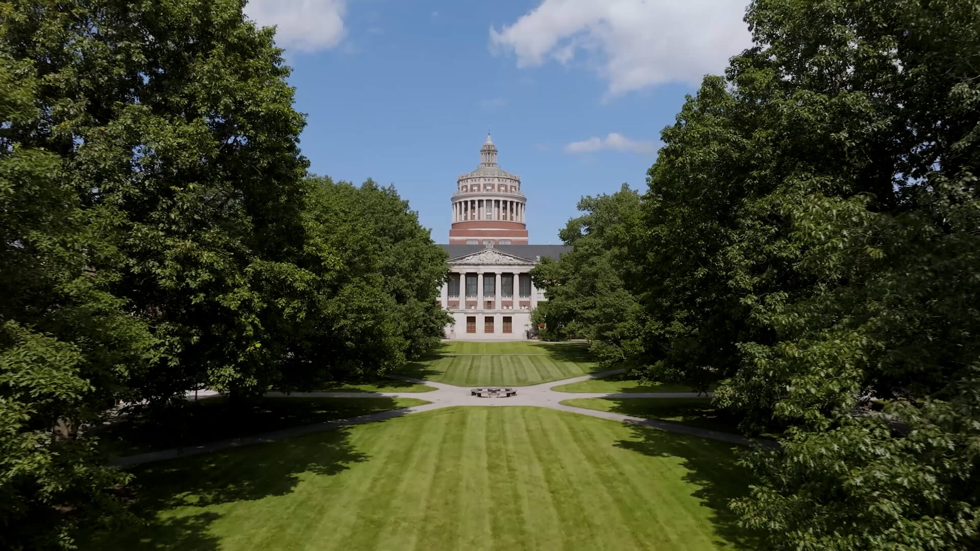 Rush Rhees Library rises between a welcoming embrace of trees, with sunlit lawns and crossing paths leading toward its doors.