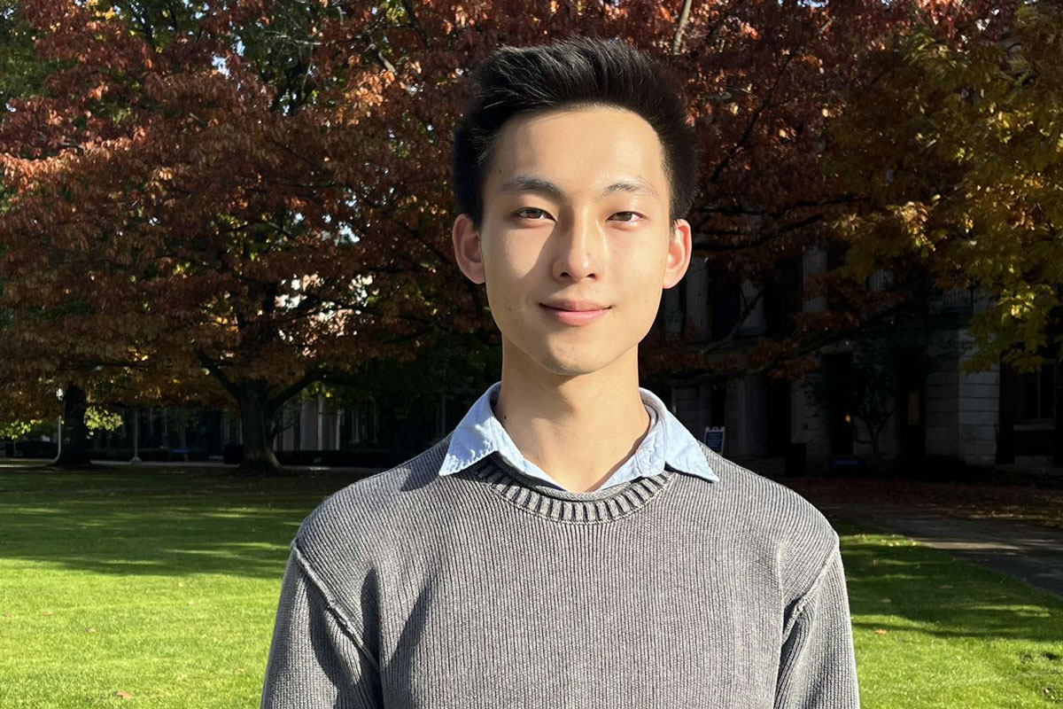 A University of Rochester student in a gray sweater stands in front of a tree, showcasing a serene campus environment.