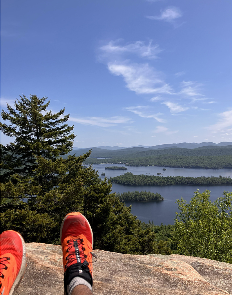 A view of wilderness in the background with legs and orange hiking shoes in the foreground.