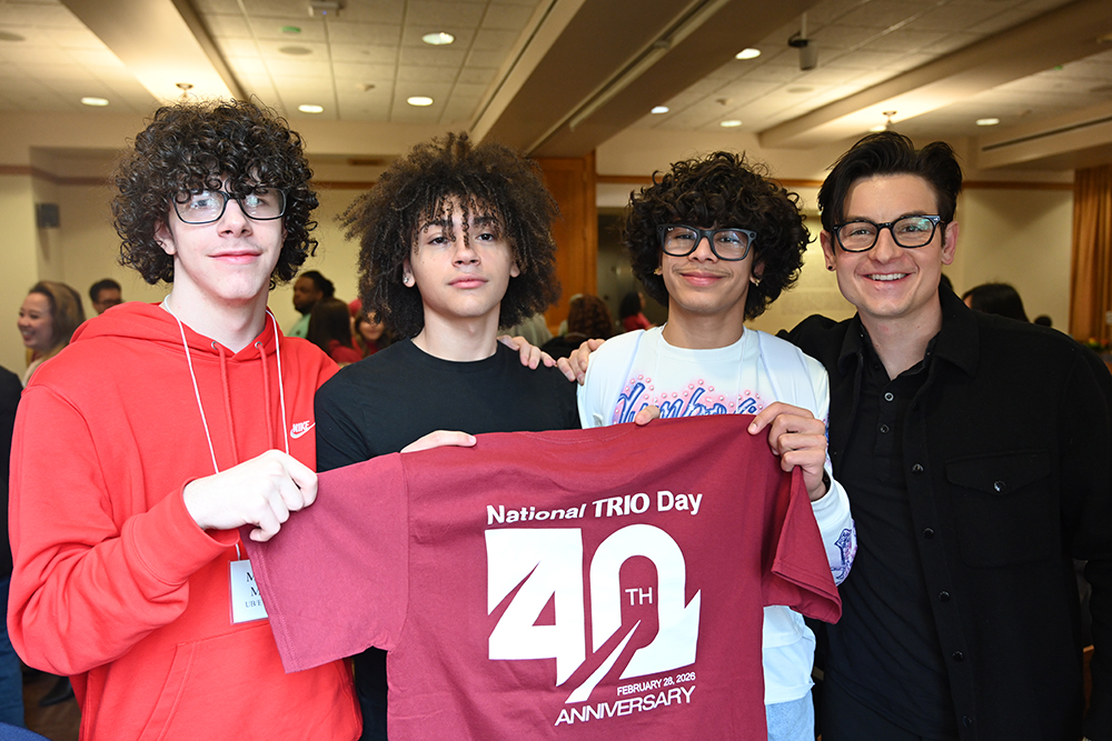 Four people posing for the camera while holing a red t-shirt with the 40th anniversary Trio Day logo.