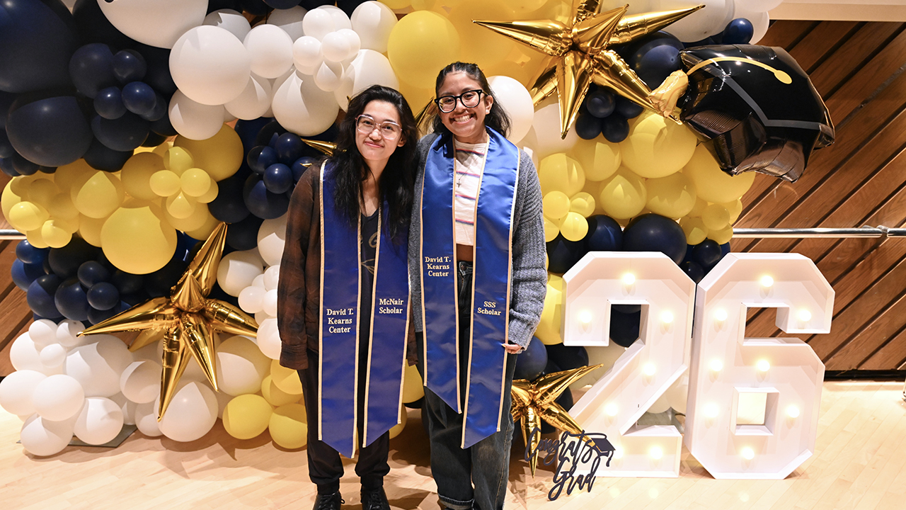 Two people wearing regalia stand in front of a blue and yellow balloon wall.