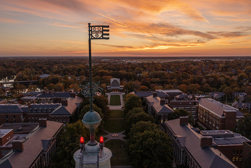 Aerial drone photo of Meliora weathervane atop Rush Rhees Library Tower, as seen during sunset.