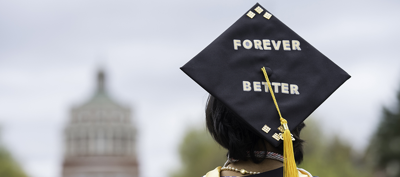 a student with a graduation cap on that reads "forever better" looking at Rush Rhees Library on commencement day