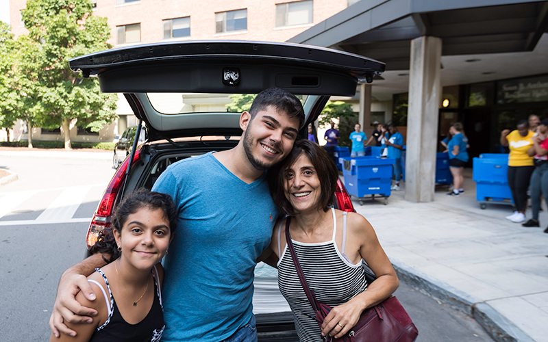 Photo of a student posing with their parent and younger sibling in front of their car outside Susan B. Anthony Residence Hall