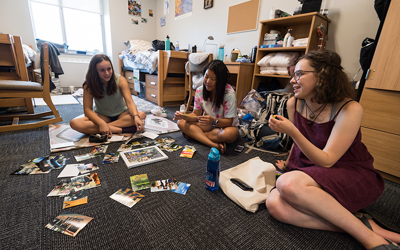 Photo of three students sitting on the floor of an on-campus dorm room talking and looking at photos