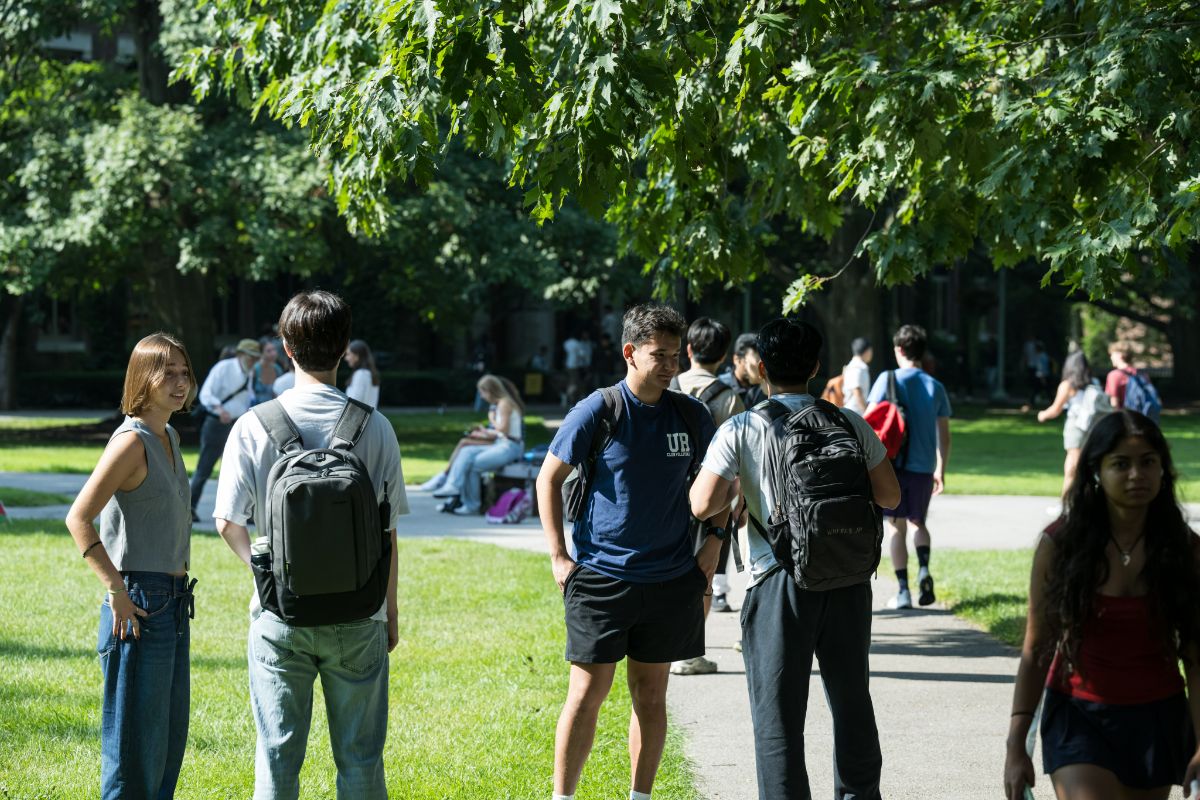 People standing and walking in a grassy outdoor area with pathways and trees, resembling a university campus.