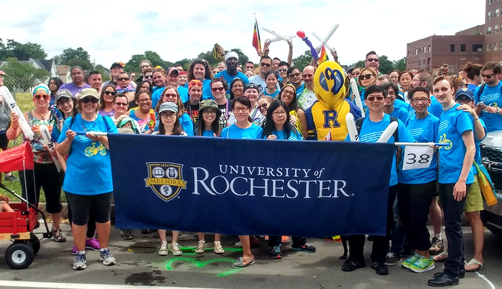 large group pf people and the Rocky yellowjacket mascot march in Pride Parade behind banner reading UNIVERSITY OF ROCHESTER