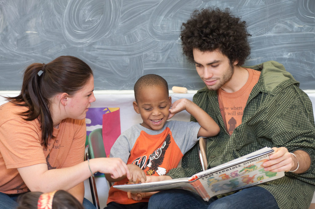 Two University of Rochester students reading a picture book to a young child in a children's classroom