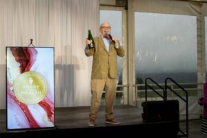 auctioneer holds open bottle of champagne on stage in front of cream backdrop