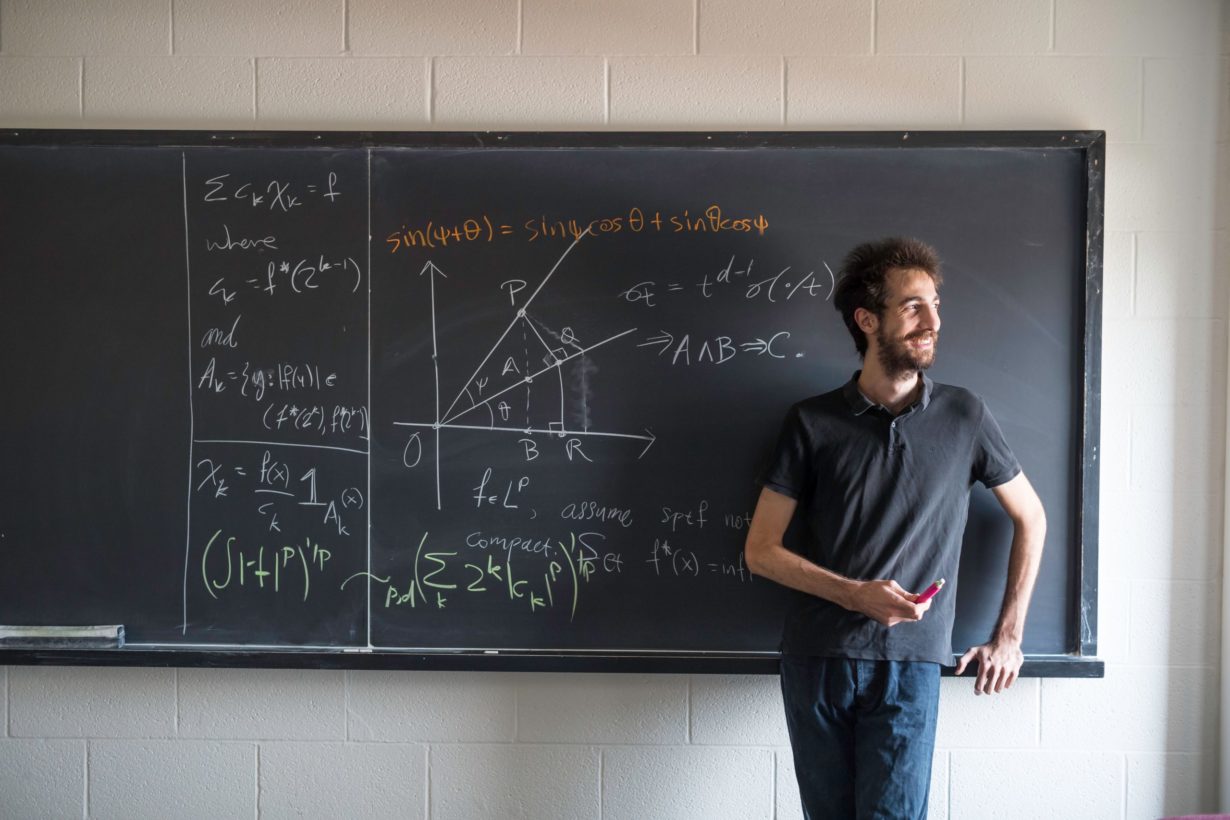 A University of Rochester graduate student in front of a chalk board with equations written all over it
