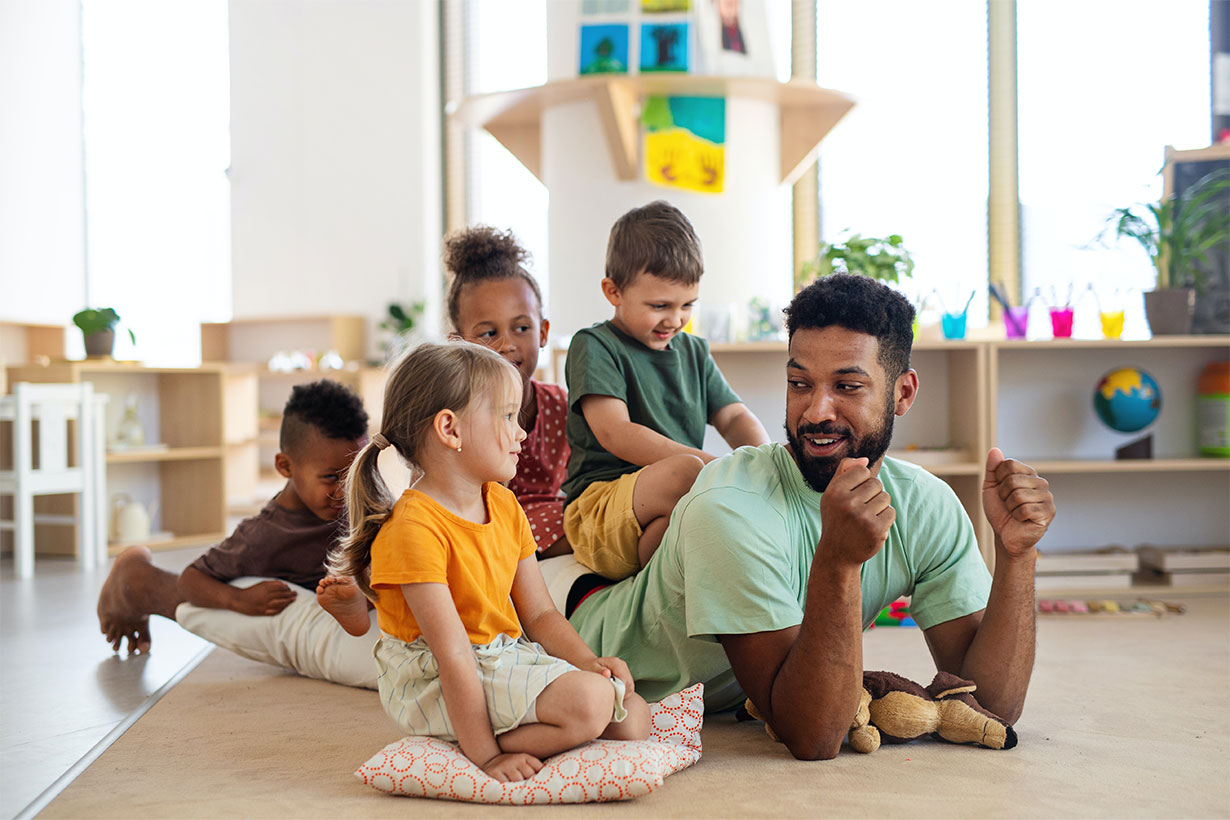 Group of small nursery school children with man teacher sitting on floor indoors in classroom, playing.