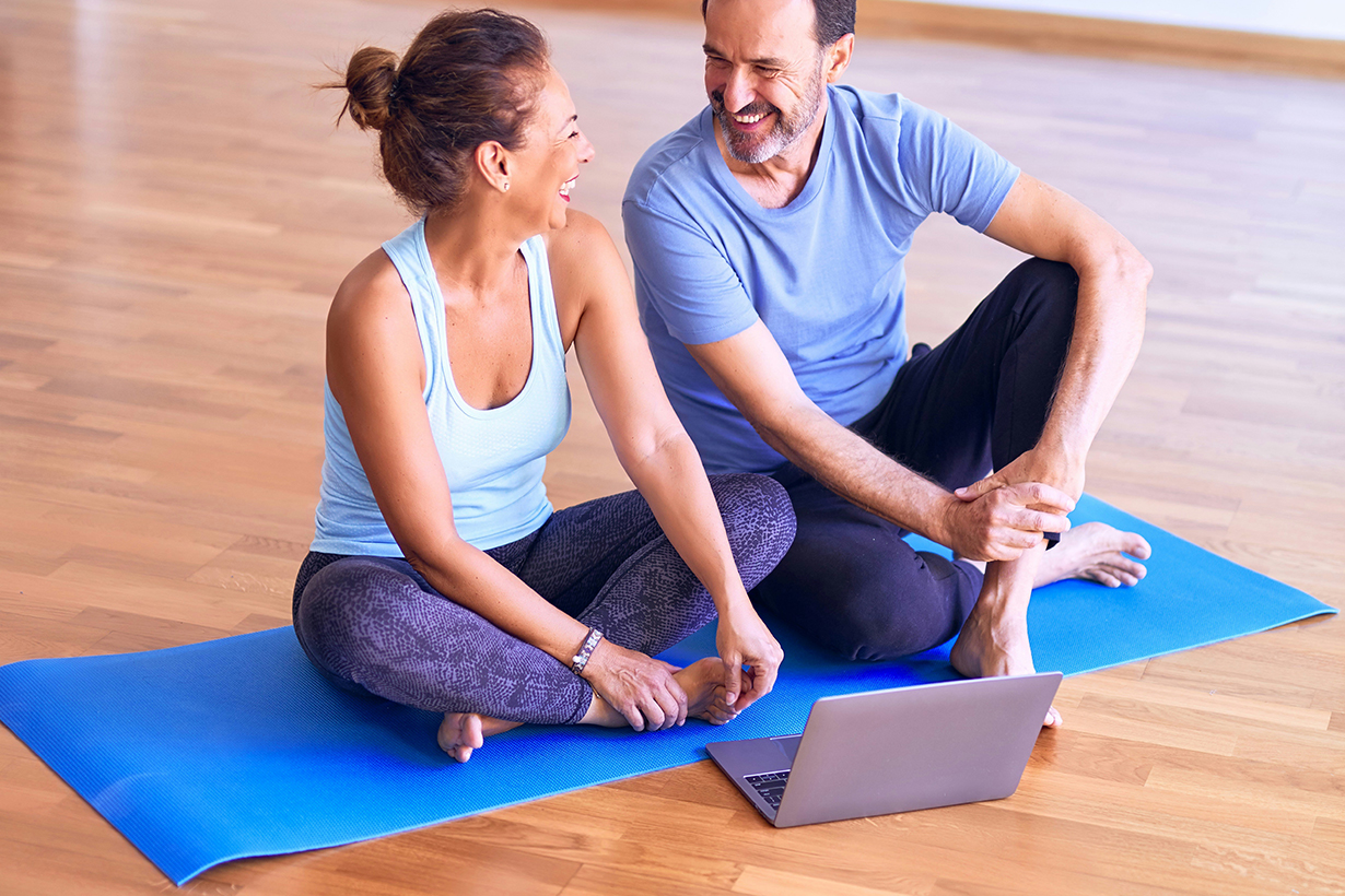 Middle age sporty couple smiling while sitting on a yoga mat with a laptop in front of them.