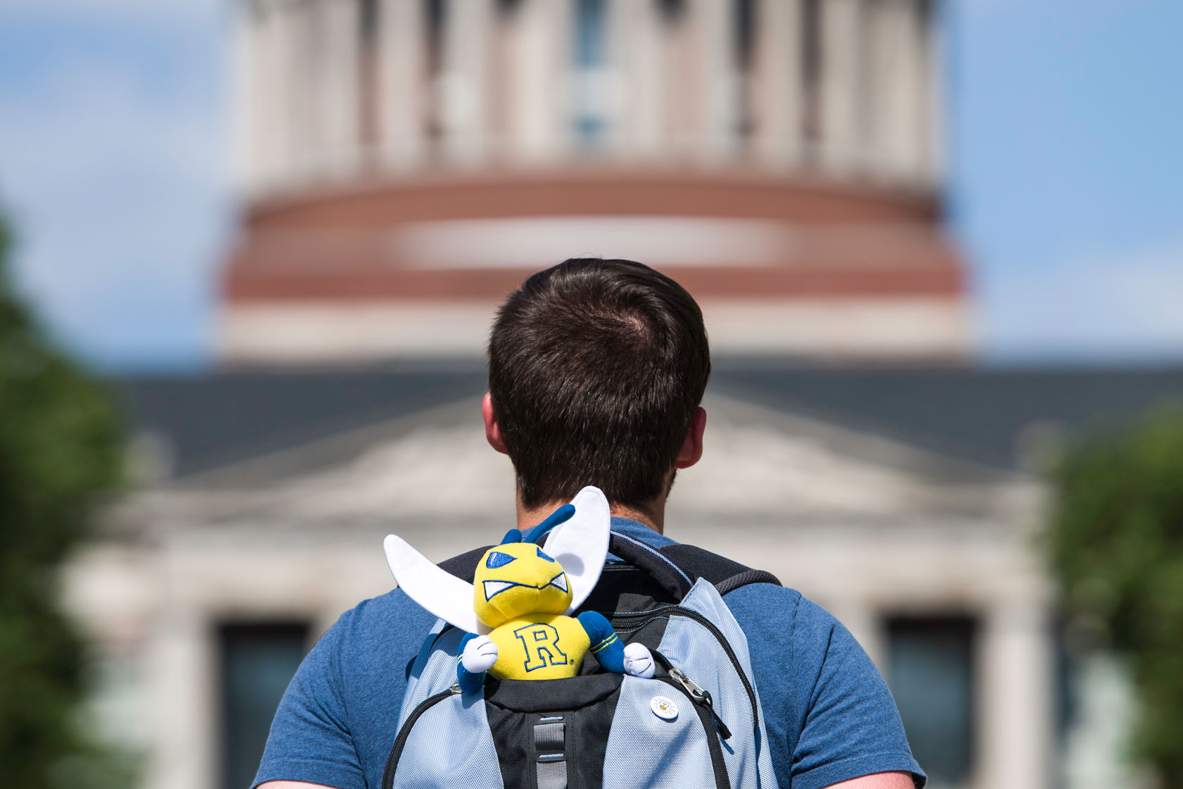 A person with short brown hair is seen from behind, wearing a blue shirt and carrying a light blue and gray backpack. Attached to the backpack is a small plush mascot with a yellow face, blue wings, and a letter 