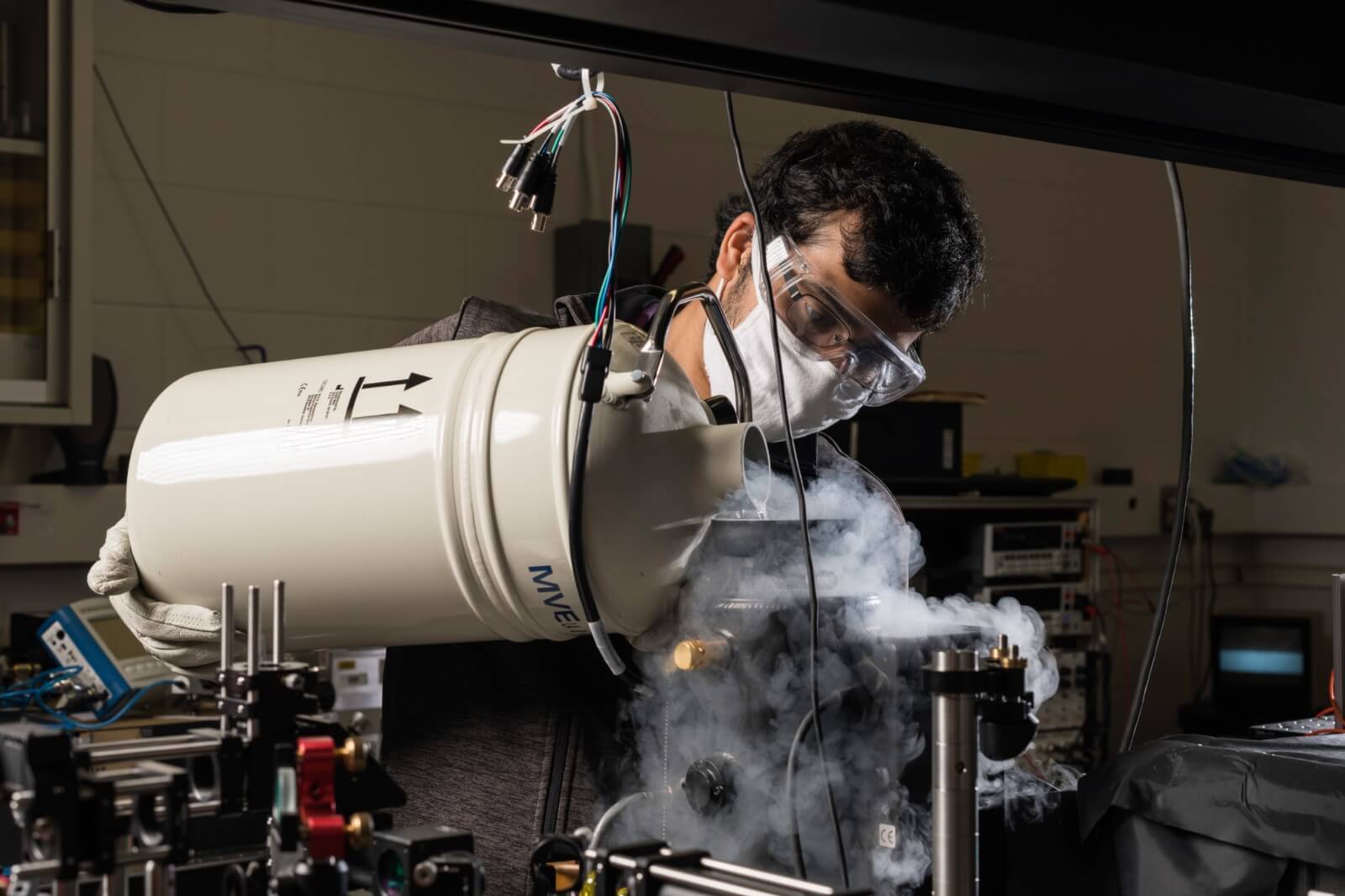 A scientist wearing safety goggles and a face mask carefully pours liquid nitrogen from a large white container into scientific equipment surrounded by various cables and devices in a laboratory setting. Cold vapor is visible emanating from the container and equipment.