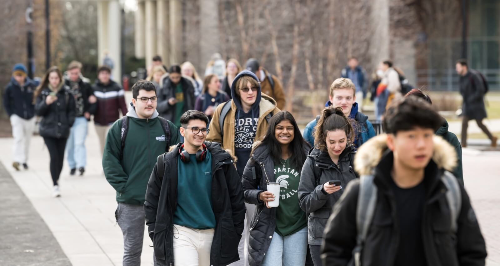 A group of college students walk along a campus pathway wearing coats and backpacks. Some are smiling and chatting, while others are looking at their phones or ahead. The background shows more students and campus buildings on a clear, cold day.