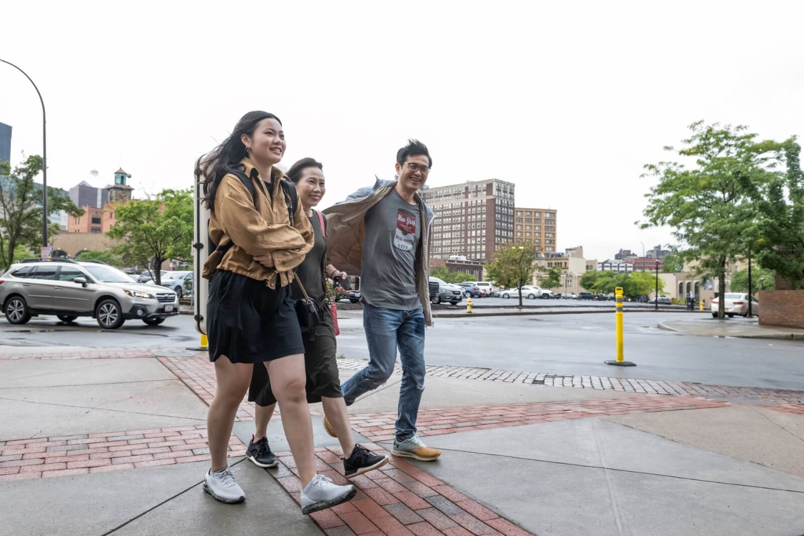 Three people walking on a sidewalk, smiling and appearing to be engaged in conversation. One person on the left wears a light brown jacket, the middle person wears a green jacket, and the person on the right wears a gray shirt and jeans. Urban background with cars and buildings.