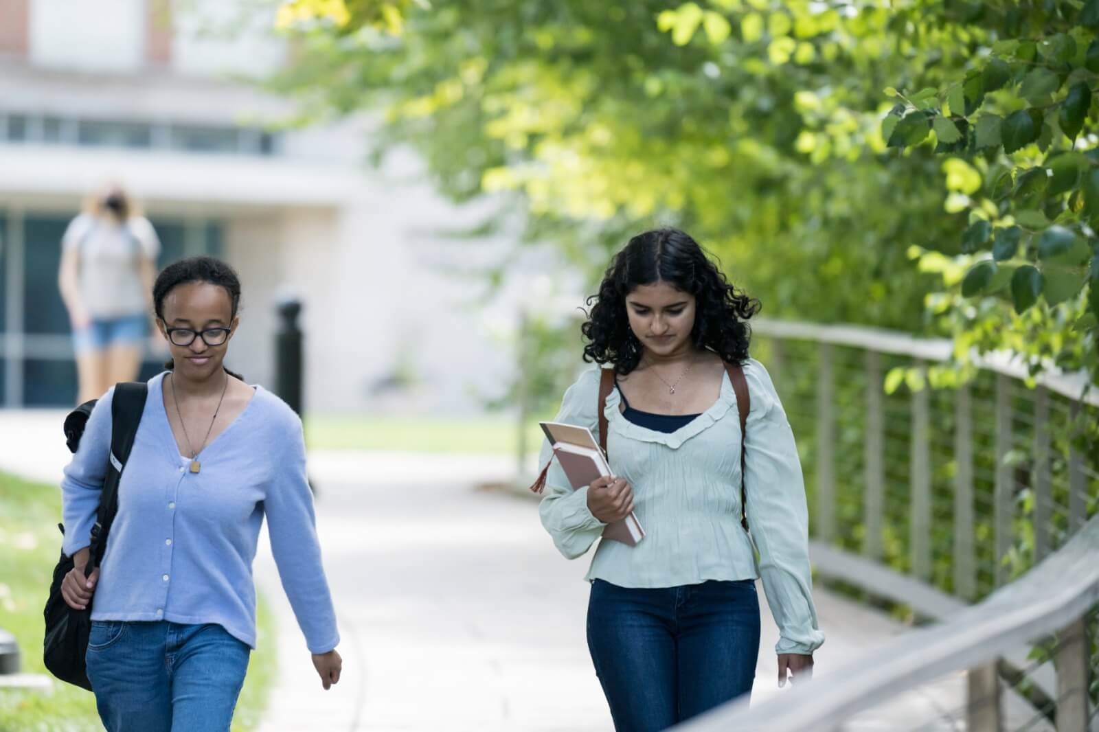 Two individuals walking along a shaded pathway lined with green trees. The person on the left wears glasses, a light blue cardigan, and carries a bag over their shoulder. The person on the right holds books and wears a light green blouse and jeans.