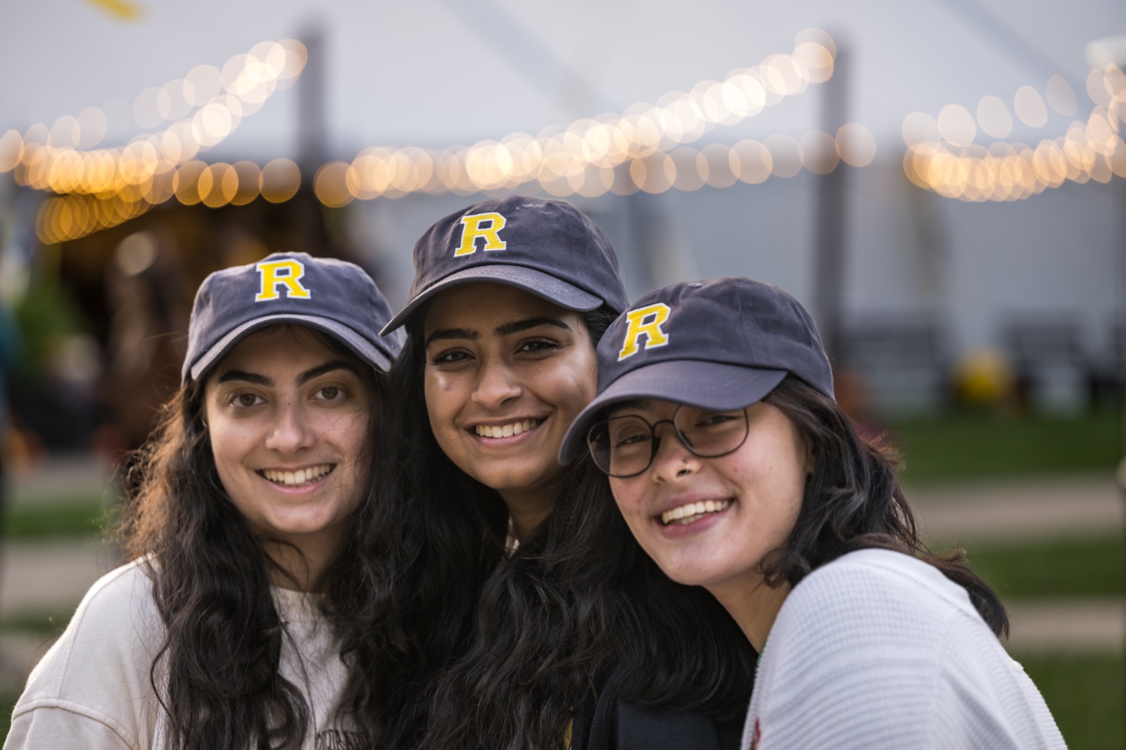 Three smiling people pose together outdoors, each wearing a navy blue cap with a yellow letter 